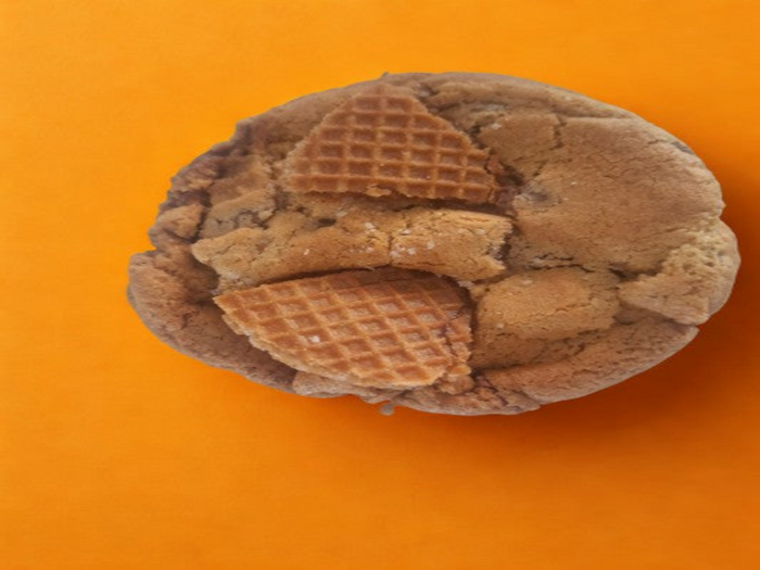 A round chocolate chip cookie with two broken waffle pieces embedded on top, photographed from above on a solid orange background.